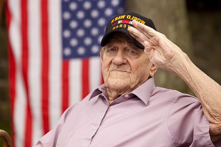 Older man wearing cap that reads "World War II Veteran" salutes with United States flag in background