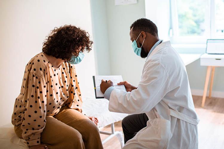 Man wearing white coat and surgical mask reviews notes with woman sitting on patient exam table wearing polka dot blouse and surgical mask